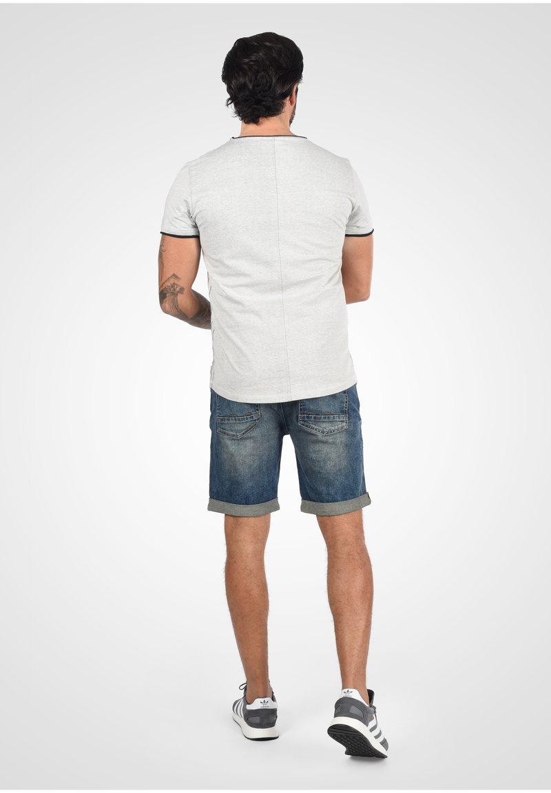 Camisa gris de manga corta con ribete negro, combinada con pantalones cortos de mezclilla azules enrollados en el dobladillo, modelo visto desde atrás, llevando zapatillas grises.