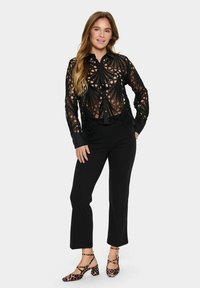 Crna prozirna čipkana bluza s složenim izrezima, uparena s uskim crnim hlačama i cipelama s blok petom u leopard printu. Duga kosa stilizirana spušteno.