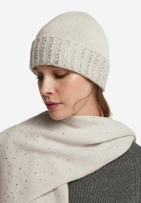 Kremowa dzianinowa czapka z prążkowanym wzorem i metalicznymi akcentami. W połączeniu z jasnym szalikiem ozdobionym srebrnymi ćwiekami na szarym topie.