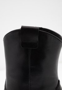 Stivaletto in pelle nera con forma arrotondata, superficie strutturata e un'etichetta a loop sul retro per facilitarne l'indossamento. Presenta cuciture visibili.