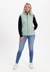 Hellgrüne gesteppte ärmellose Weste mit hohem Kragen, Frontreißverschluss und seitlichen Taschen, getragen über einem schwarzen Langarmoberteil und blauen Jeans.