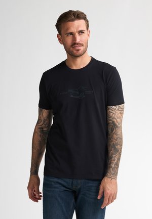 Zwarte T-shirt met een grafisch ontwerp van een auto in donkere print. Korte mouwen en een ronde halslijn. Gecombineerd met denim jeans.