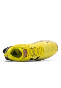 Zapatilla de running amarilla con upper de malla, acentos negros y suela texturizada. Presenta cordones prominentes y una marca en la lengüeta.
