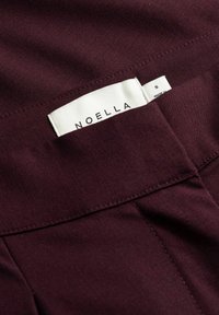 Λευκή ετικέτα ρούχου με "NOELLA" και μέγεθος "S" τοποθετημένη μέσα στη ραφή ρούχου από σκουρόμωβ ύφασμα.