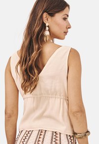Top sin mangas de color beige claro con escote en V y cintura fruncida. Tejido texturizado, combinado con una falda de estilo bohemio con estampados y joyería decorativa.