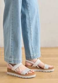 White woven sandals feature a cork and white platform, with adjustable straps and a textured finish, worn with light blue jeans.