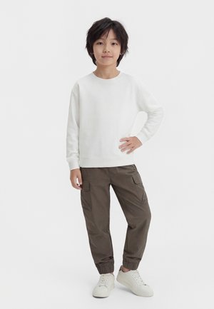 Chlapec stojaci s jednou rukou na bok, oblečený v bielej dlhorukávovej košeli, hnedých cargo nohaviciach a bielych teniskách na jednolôžkovom pozadí.