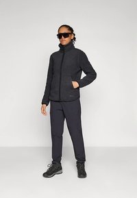 Jachetă din fleece gri închis cu guler înalt, închidere cu fermoar și buzunare laterale. Asortată cu pantaloni închiși la culoare și bocanci negri de drumeție.