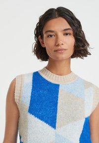 Knit sweater with a geometric pattern in blue, light blue, cream, and beige. Features a ribbed neck and sleeveless design. Soft texture.