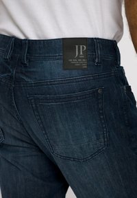 Unos pantalones vaqueros de denim azul oscuro presentan un parche de cuero en la parte trasera, bolsillos cosidos y un corte ajustado, resaltando una textura suave.