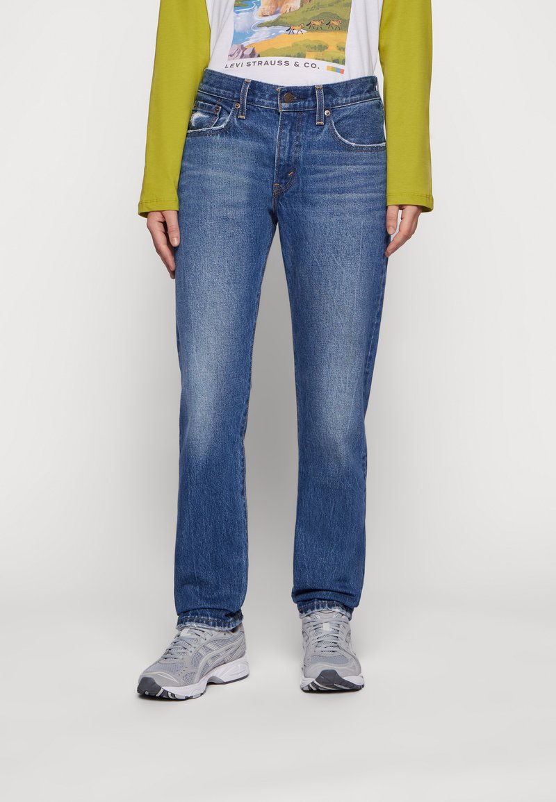 Jeans de mezclilla azules con corte recto, ligero desgaste, cinco bolsillos y cierre de botón. Combinados con zapatillas deportivas grises y una camiseta gráfica.