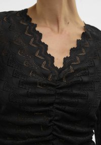 Schwarzes Spitzenoberteil mit einem gewellten Ausschnitt, strukturierten Rüschendetails und einem kunstvollen Muster im gesamten Stoff.