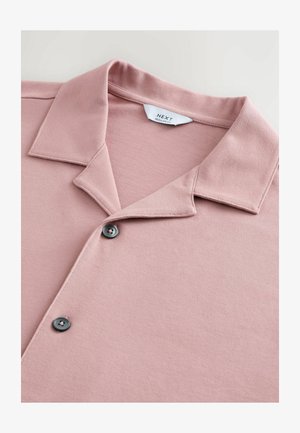 Roza polo majica izrađena od teksturirane tkanine, s kontrastnim dugmetom i klasičnim dizajnom ovratnika. Oznaka vidljiva unutar ovratnika.