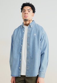 Lichtblauwe denimhemel met knopen aan de voorkant, lange mouwen en een klein logo op de linker borst. Draagt over een wit t-shirt en groene broek.