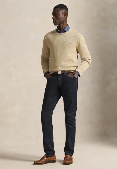 Beige strikkegenser, blå rutete skjorte under, mørke denimjeans, brune skinnsko, med en klokke på venstre håndledd.