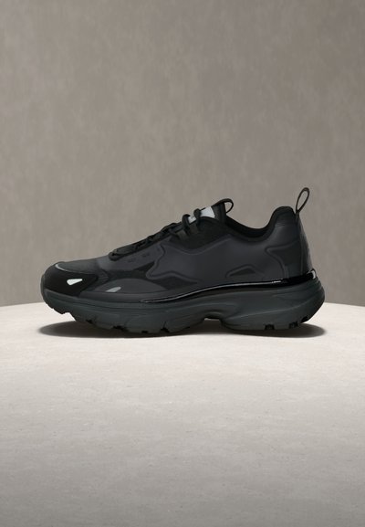 Baskets noires à semelle épaisse, design à lacets et languette de traction sur fond neutre, vue de côté.