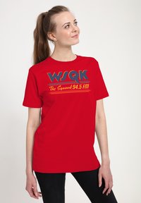 Kvinde med langt hestehale iført en rød T-shirt med "WSQK The Squawk 94.5 FM" logo, der kigger opad og stående mod en ensfarvet baggrund.