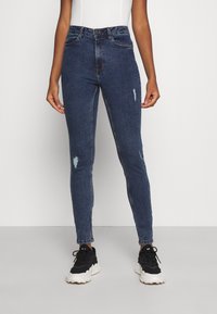 Tmavě modré vysoké skinny džíny s oprýskanými detaily, pět kapsami a lehkým vyblednutím na nohavicích. Vytvořeno s černými teniskami.