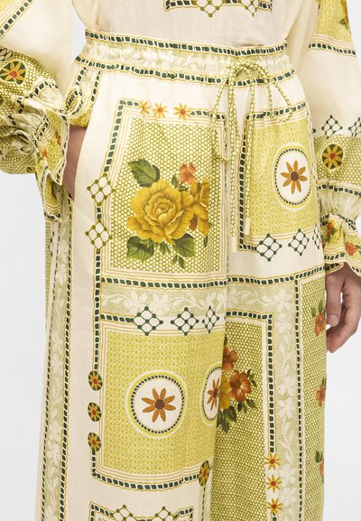 Χαλαρό παντελόνι με πατς από λουλουδάτα μοτίβα σε κίτρινο και πράσινο, με μεγάλα κίτρινα λουλούδια και ελαστική μέση με κορδόνι. Τα χέρια βρίσκονται στις τσέπες.