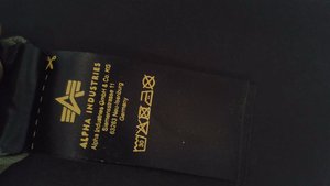 Mustast kangast hooldusjuhendi silt kollase tekstiga, mis kuvab brändi nime "Alpha Industries", hooldusjuhised ja tootmisteave.