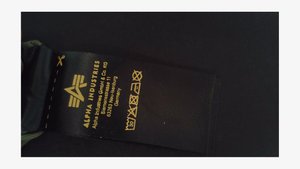 Melna auduma kopšanas etiķete ar dzeltenu tekstu, kas attēlo zīmola nosaukumu "Alpha Industries", kopšanas instrukcijas un ražošanas informāciju.