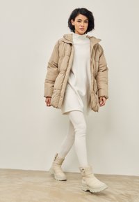 Veste en duvet beige avec capuche, tunique blanche et leggings blancs. Bottes chunky beiges avec fermetures éclair et semelles texturées. Design simple et moderne.