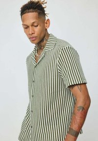 Chemise à manches courtes avec boutons, présentant un motif rayé vert et blanc, un design col, et un tissu texturé.