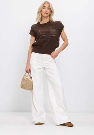 Blonde vrouw draagt een lichtbruine, gebreide top met korte mouwen, witte brede broekspijpen, beige schoenen en houdt een beige gevlochten handtas vast.