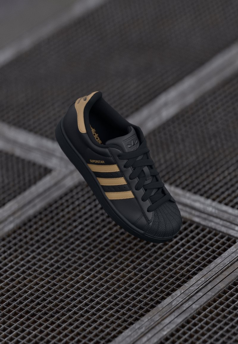 Black sneaker with three gold stripes, textured shell toe, and lace-up design. Features brand logo on tongue and side.