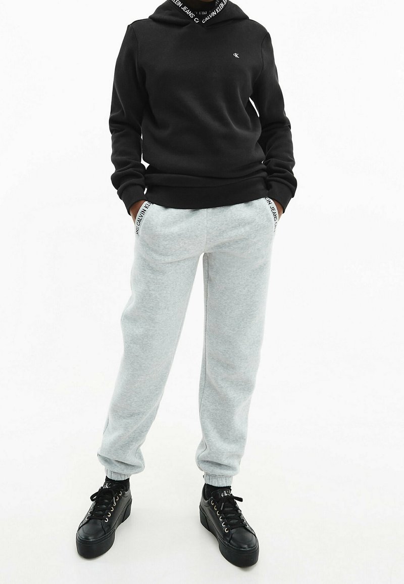 Felpa nera con un logo sul petto, abbinata a pantaloni della tuta grigi con accenti del logo sulle tasche e scarpe nere con lacci.