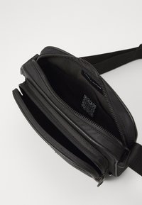 Schwarze Nylon-Crossbody-Tasche mit zwei Reißverschlussfächern, glatter Textur und verstellbarem Tragegurt. Enthält ein Innenlabel und einen QR-Code.