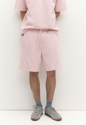 Muž v svetloružových šortkách a tričku, sivé pásikavé ponožky a sivé semišové tenisky, stojaci s rukami za chrbtom.