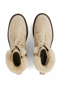 Botas de tobillo de ante beige con un forro esponjoso de color crema, detalles en dorado y cordones estándar. Suelas de goma marrón gruesa que ofrecen tracción.