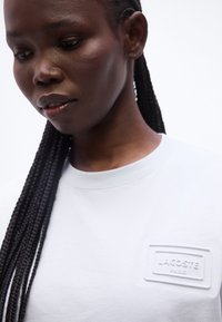 Κοντινό πλάνο ενός ατόμου με μακριά πλεξουδωτά μαλλιά που φοράει λευκό πουκάμισο με στρογγυλή λαιμόκοψη, με ένα ανάγλυφο έμβλημα λογότυπου Lacoste Paris.