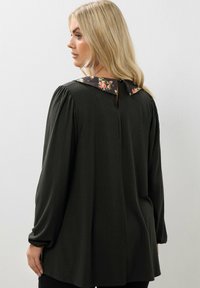 Donna con lunghi capelli biondi, che indossa una maglietta nera a maniche lunghe con colletto floreale, vista di spalle su uno sfondo bianco.