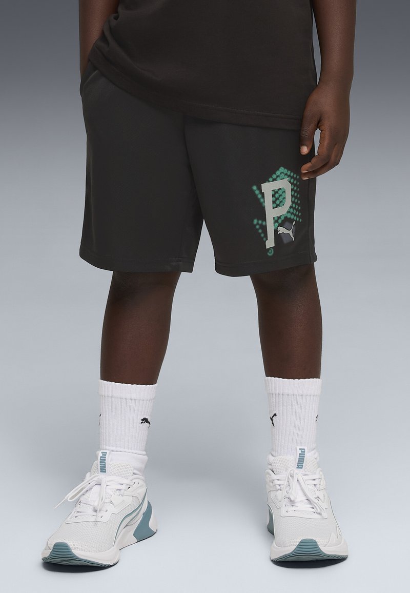Enfant portant un short noir Puma avec un logo "P" turquoise, des chaussettes blanches Puma et des chaussures de sport blanches avec des accents turquoises, debout sur un sol gris.