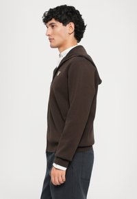 Brown zip-up hoodie with a hood, featuring ribbed cuffs and hem. Worn over a collared white shirt. Smooth texture and fitted design.