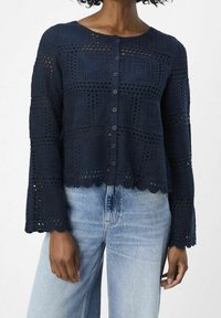 Cárdigan de crochet marino con un patrón cuadrado texturizado, dobladillo con ondas y cierre de botones. Combinado con jeans de color azul claro.