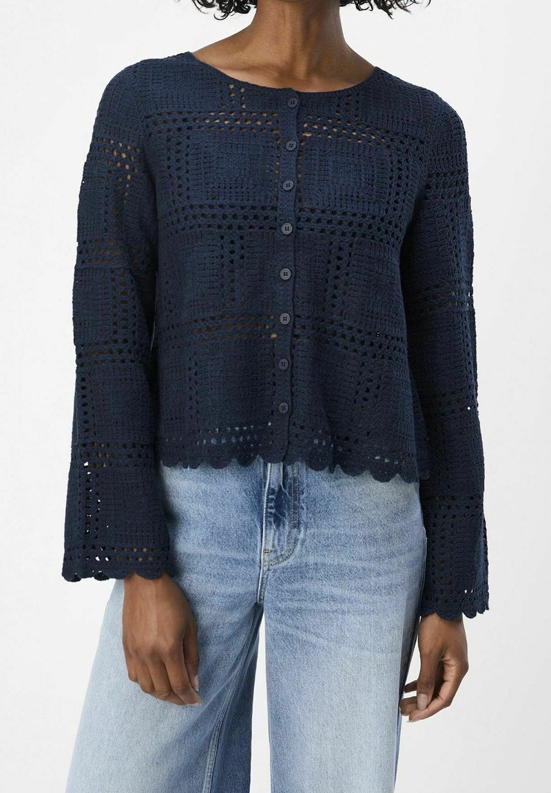 Cárdigan de crochet marino con un patrón cuadrado texturizado, dobladillo con ondas y cierre de botones. Combinado con jeans de color azul claro.