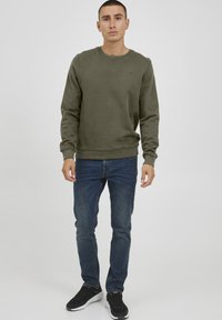 Sudadera de cuello redondo en verde oliva, fabricada en un tejido suave, combinada con unos jeans de color azul oscuro y zapatillas negras. Diseño sencillo con puños y bajo acanalados.