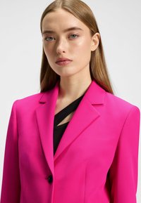 Blazer sur-mesure fuchsia avec des revers marqués et une fermeture à un bouton, porté sur un haut noir avec un design découpé. Texture de tissu lisse.