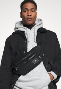 Bolso bandolera negro hecho de tela duradera, con un bolsillo con cremallera y un parche de logo. Modelo vistiendo una sudadera gris y una chaqueta negra.