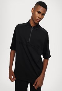 Svart kortermet polo skjorte med glidelåskrage, med en teksturert logo i midten og et jevnt, stretchy stoff.