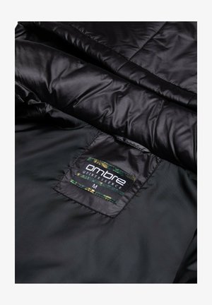 Chaqueta acolchada negra con una textura suave, presenta una etiqueta negra cosida en el interior que dice "esencia ombré" y tamaño "M".