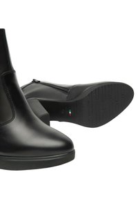 Stivali in pelle nera alla caviglia con tacco a blocco, zip laterali e suole in gomma testurizzata; uno stivale in posizione verticale e l'altro disteso di lato.