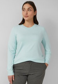 Vrouw met donker haar draagt een lichtblauwe langslapige trui en grijze broek, staat met één hand in de zak en kijkt opzij.
