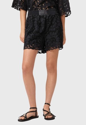Sorte blonder shorts med blomsterprint og bølgekant, båret med sorte flade sandaler med stropper på en model, der står mod en lys baggrund.