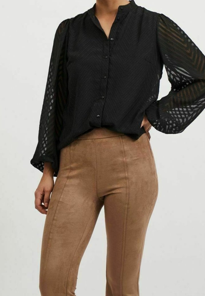 Blouse noire à boutons avec manches transparentes texturées ; associée à un pantalon moulant en faux daim marron clair avec des coutures verticales.