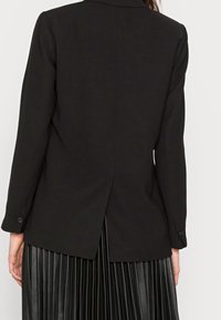 Widok z tyłu osoby w czarnym żakiecie z centralnym rozcięciem z tyłu i czarnej plisowanej spódnicy z sztucznej skóry.