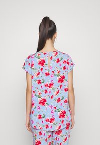 Una donna con una lunga coda di cavallo indossa un top blu e pantaloni con un motivo floreale rosso e rosa, girata di spalle su uno sfondo bianco semplice.